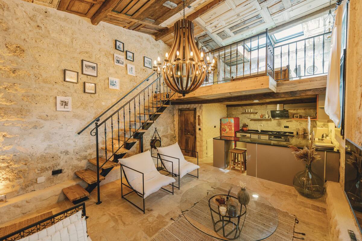 A spacious loft with a high ceiling, exposed wooden beams, a staircase with a wrought iron railing, and a kitchen area with modern appliances.