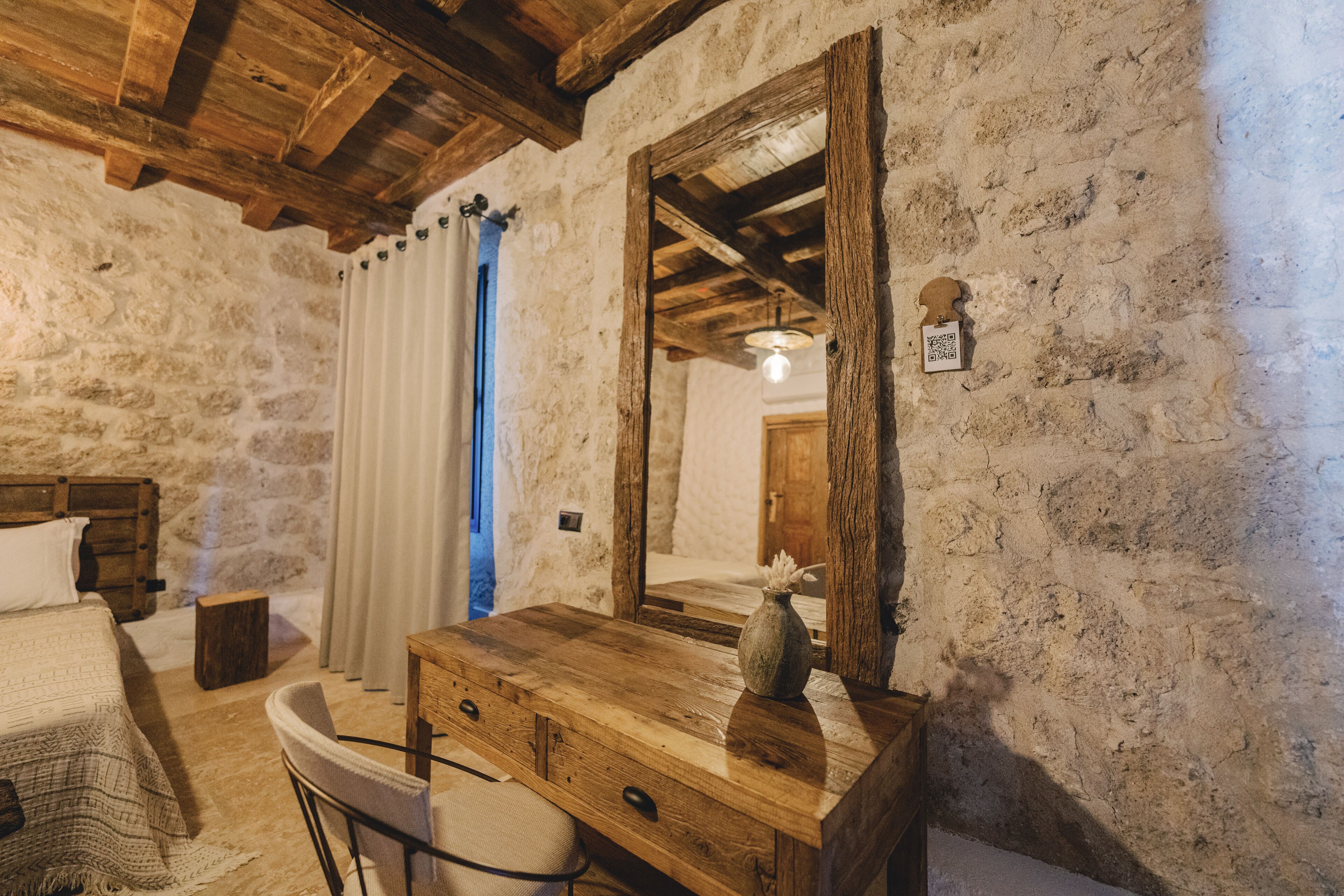 A rustic room with a stone wall, wooden ceiling beams, a bed with a white comforter, a wooden desk with a vase, and a mirror.