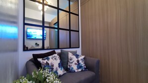 A modern living room with a grey sofa, floral cushions, a large mirror, and a wall-mounted TV. - 1BR Studio w/ Pool, Balcony, Near Airport&Hotspots (Taguig)