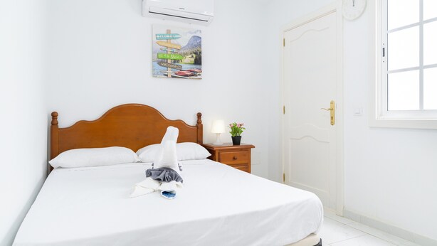 Una cama bien hecha con cabecera de madera, una mesita de noche con lámpara y una planta pequeña, un aire acondicionado instalado en la pared y una puerta con cerradura.