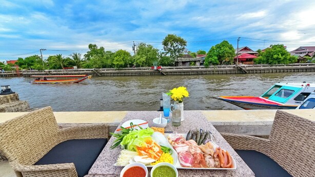 A table with a variety of food items and drinks, overlooking a river with boats and greenery in the background. - NORN Rimklong Bangkok Hotel (Bangkok)