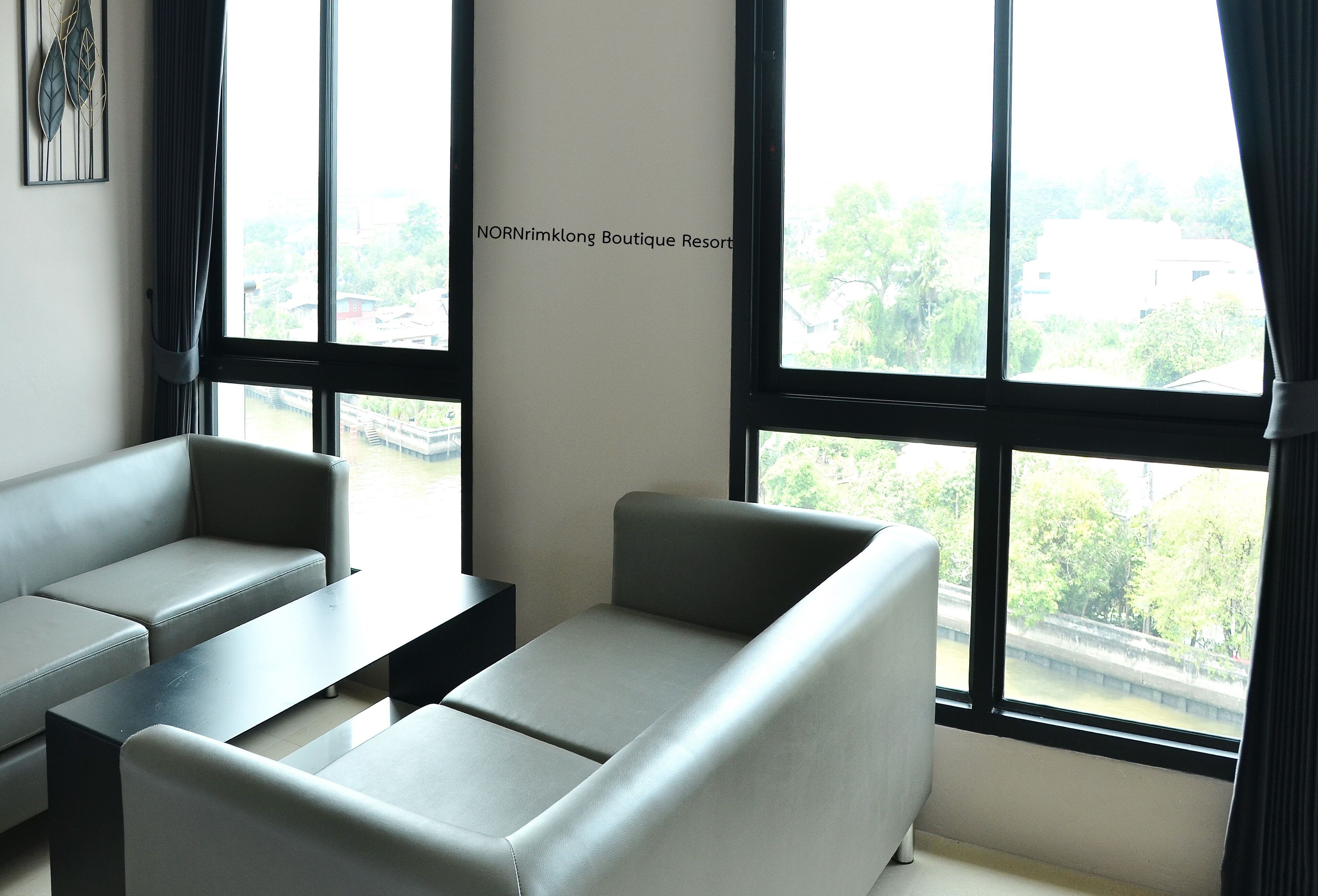 A modern room with grey sofas and a black table, large windows with curtains, and a wall sign reading "NORNrimklong Boutique Resort".