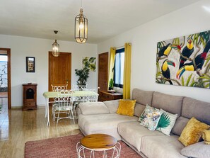 Un salon comprenant un canapé, une table à manger avec des chaises, un meuble en bois et un tableau représentant des toucans accroché au mur.