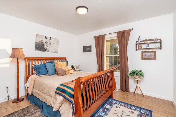 A bedroom with a wooden bed, a bedside lamp, a rug, a window with curtains, and wall decorations. - Private home on 40 acres surrounded by wide open space. (Vernon)