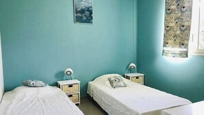 Une chambre avec deux lits simples, chacun doté d’une table de chevet et d’une lampe, d’une petite étagère fixée au mur et d’une fenêtre avec des rideaux à motifs.