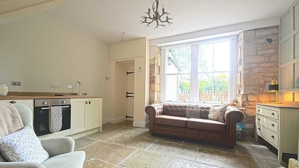 A cozy living room with a stone wall, a large window, and a built-in oven. - Coquetdale Cottage in Rothbury, quiet village location (Rothbury)