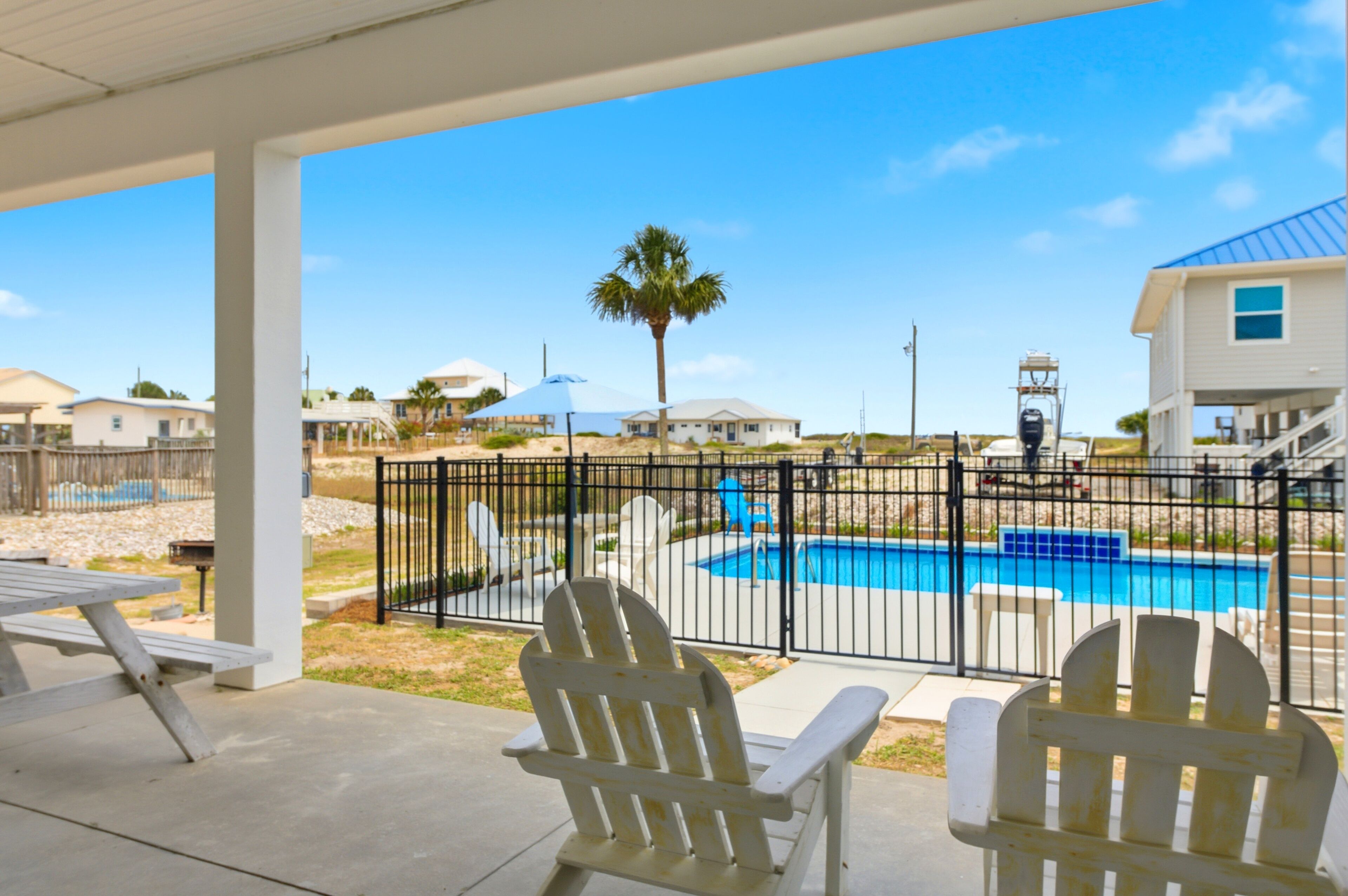 A covered patio with Adirondack chairs, a pool, and a view of houses and palm trees.