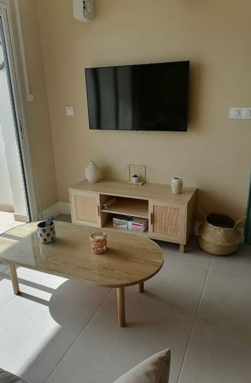 A living room with a flat-screen TV mounted on the wall, a wooden cabinet with decorative items, a glass coffee table, and a wicker basket.