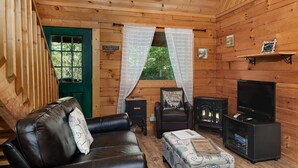 A cozy cabin interior with a leather sofa, a patterned ottoman, a television, a fireplace, and a green door.