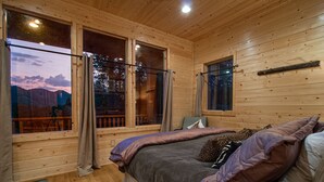 A wooden cabin bedroom with a bed, pillows, and a window overlooking a mountainous landscape.