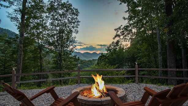 A fire pit with three wooden chairs around it, set on a gravel area with a wooden fence and trees in the background.