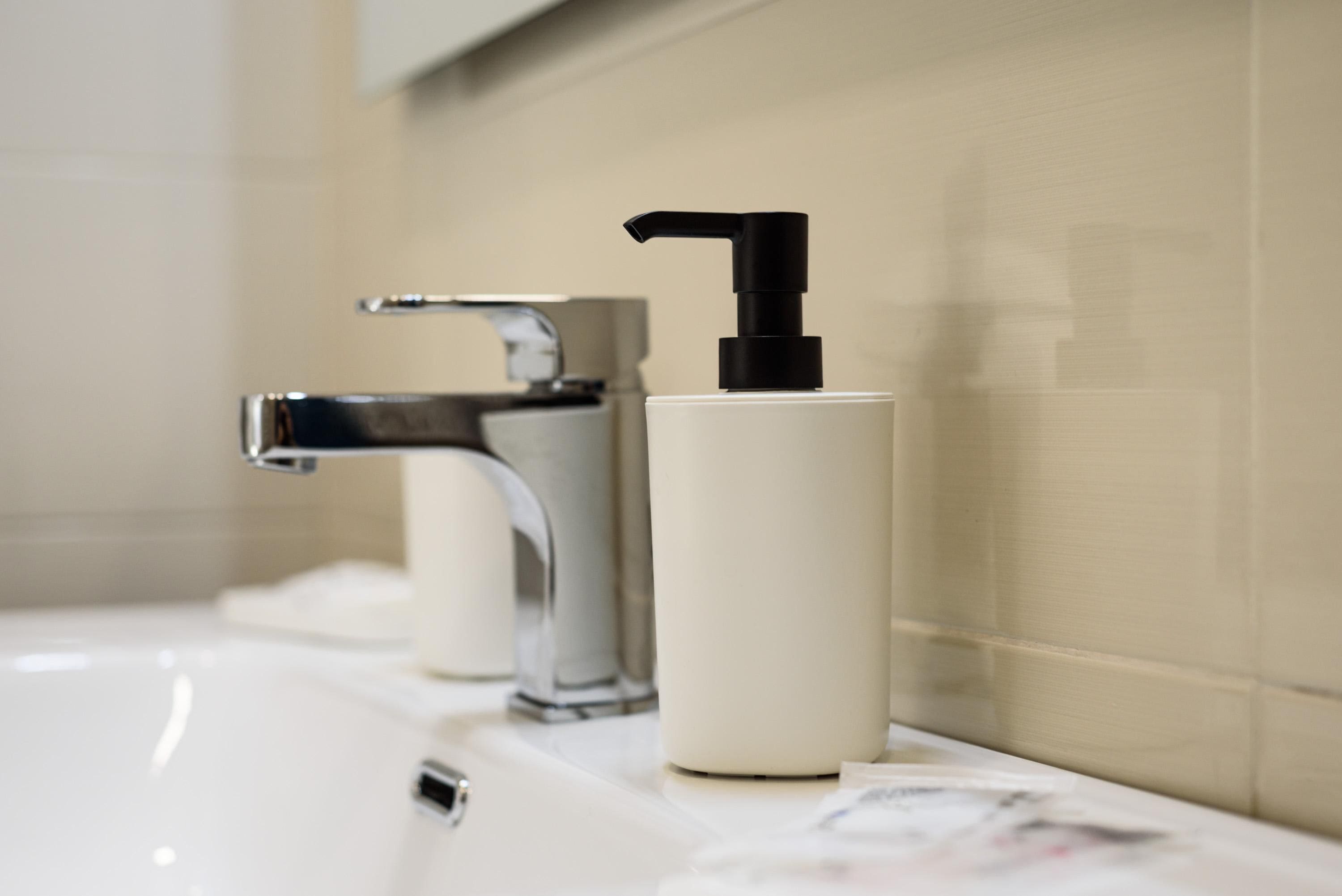Une salle de bain avec un lavabo blanc, un robinet chromé et une bouteille blanche dotée d’un distributeur noir.