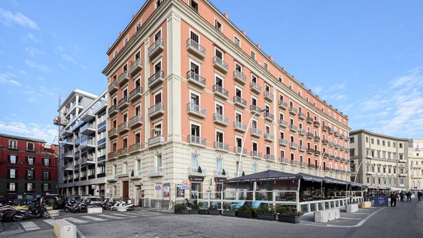 A multi-story hotel with balconies, a covered outdoor seating area, and a street in front.