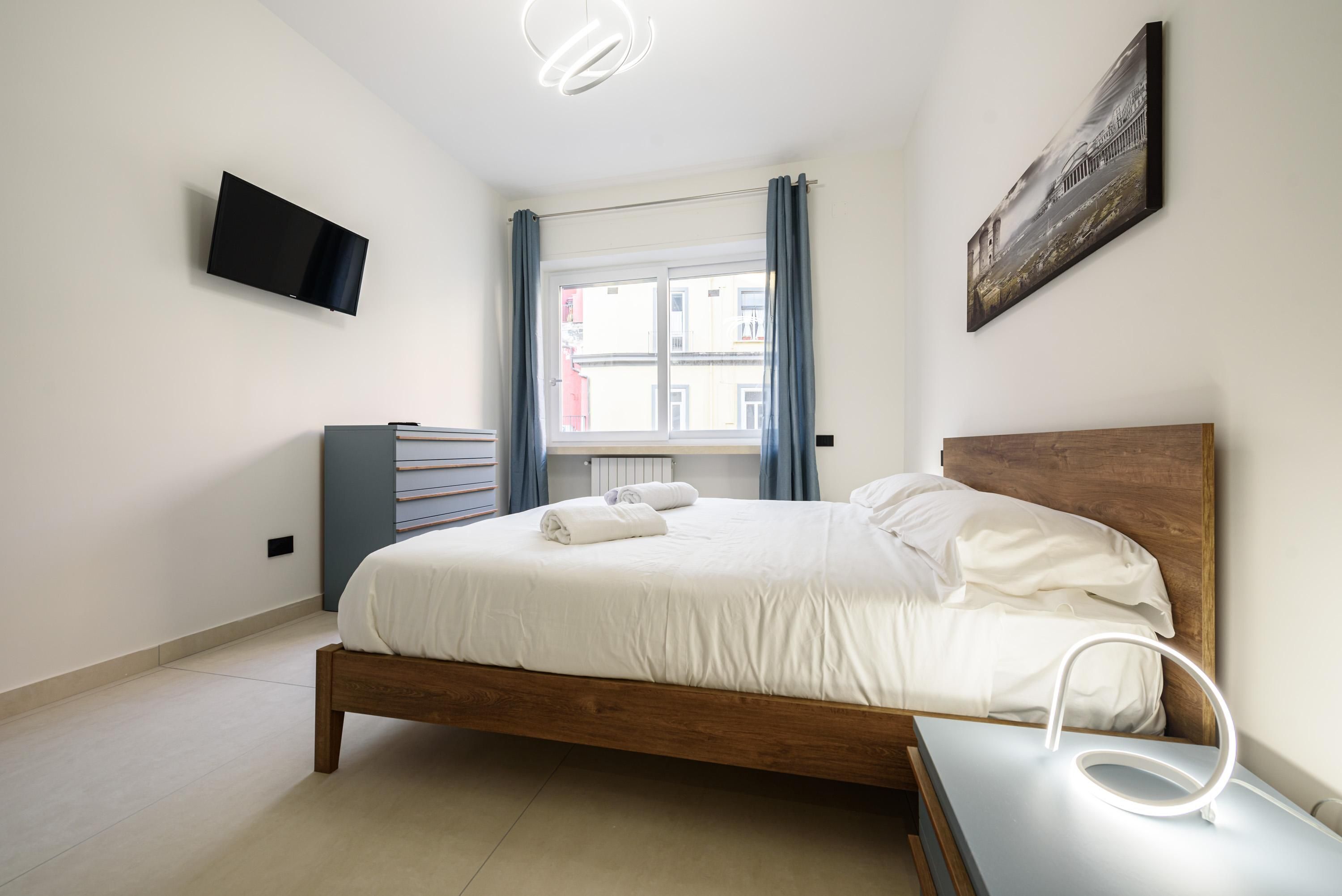 Une chambre à coucher avec un lit, un téléviseur fixé au mur, une fenêtre avec des rideaux et un tableau.