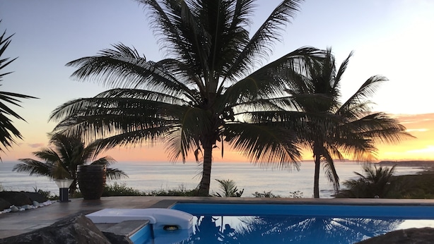 En swimmingpool med udsigt over havet i solnedgang, omgivet af palmer og klipper.