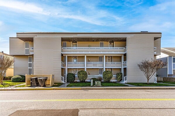 Un edificio de apartamentos de mediana altura, de dos pisos, con fachada plana, varios balcones y un pequeño jardín frontal con jardinería.