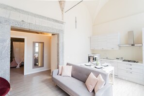 Ein modernes Wohnzimmer mit grauem Sofa, einem kleinen Esstisch und einer Küchenzeile mit weißen Schränken und einem Gasherd.