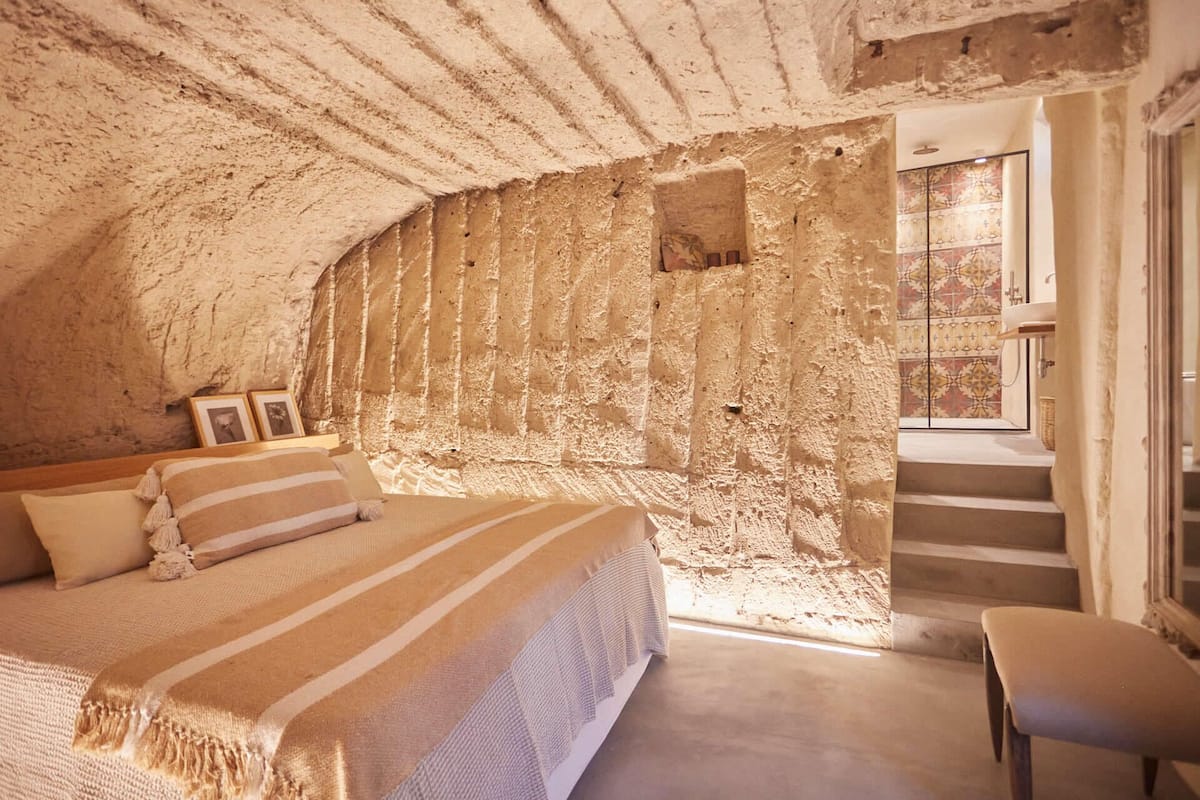 A bedroom with a stone wall, a bed with striped bedding, a chair, and a staircase leading to a bathroom.