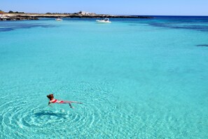 A clear blue sea with a person swimming. There are boats and buildings in the background.
