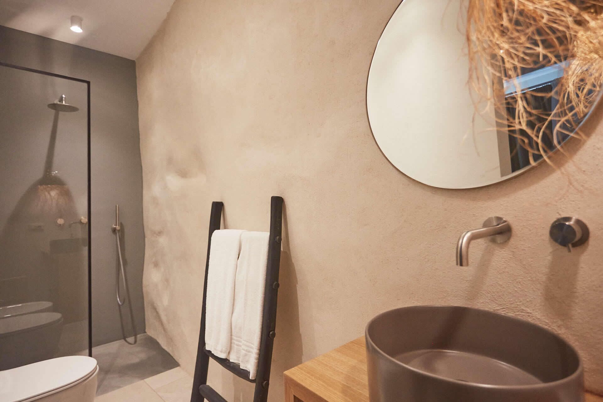 A bathroom with a glass shower enclosure, a wooden vanity with a large basin, a towel on a chair, and a round mirror on the wall.