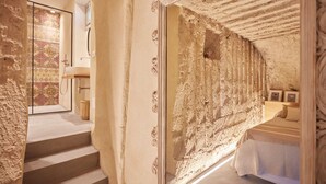 Quarto com parede de pedra rústica, cama com manta listrada e escada para um banheiro com área de chuveiro revestida em azulejo.