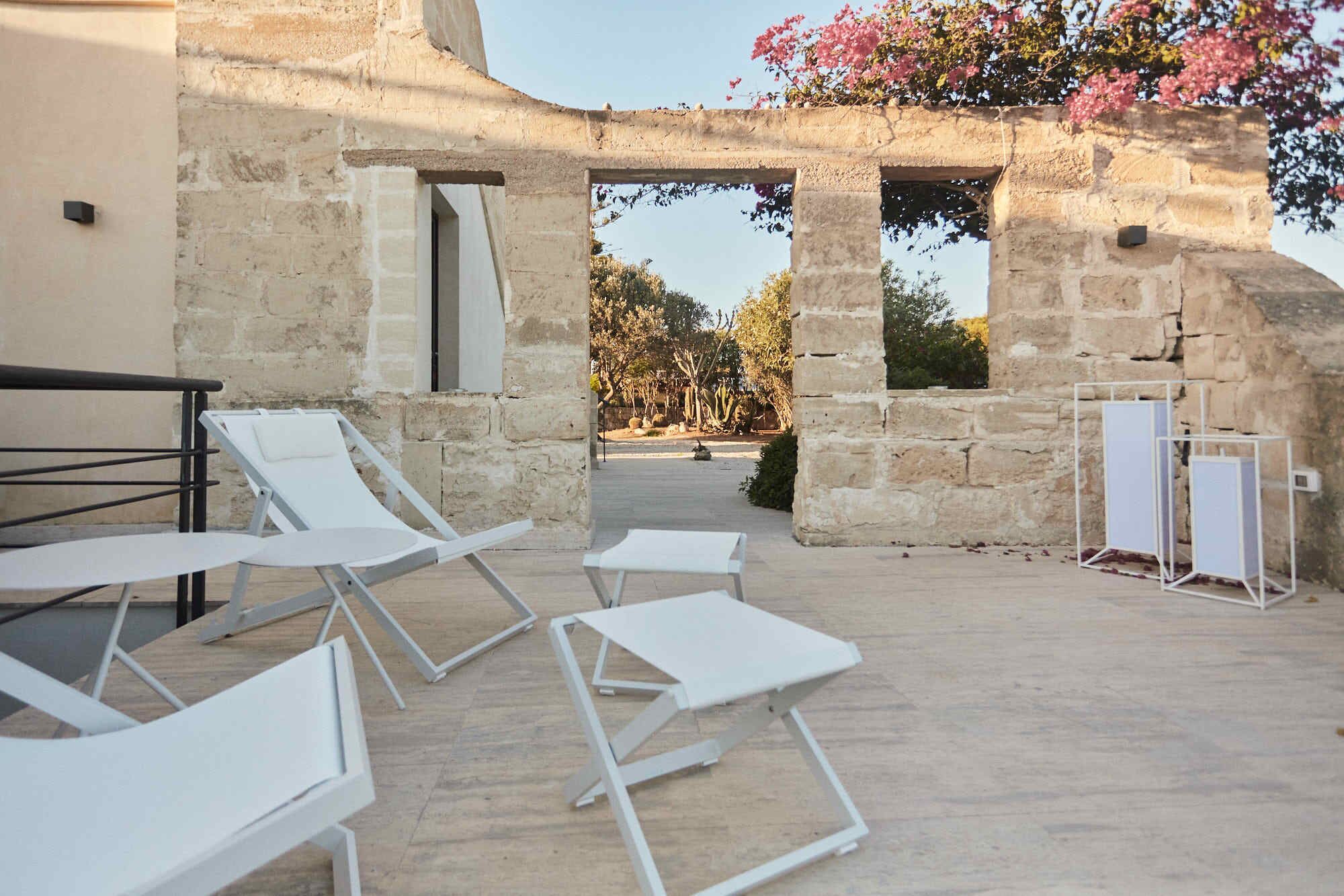 An outdoor seating area with white lounge chairs and a white table, set against a stone building with an arched doorway.