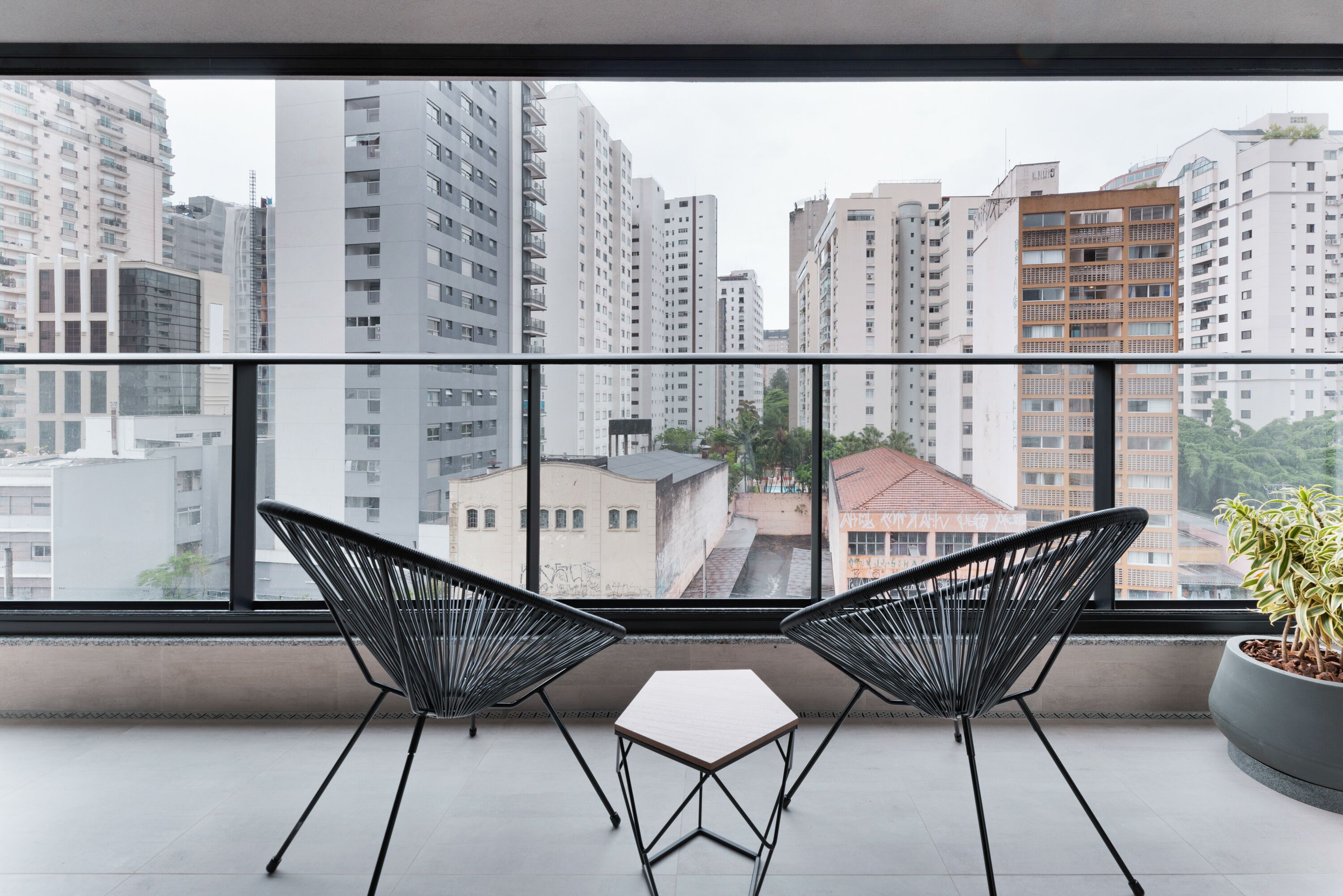 A balcony with two black wire chairs and a small table, overlooking a cityscape with tall buildings.