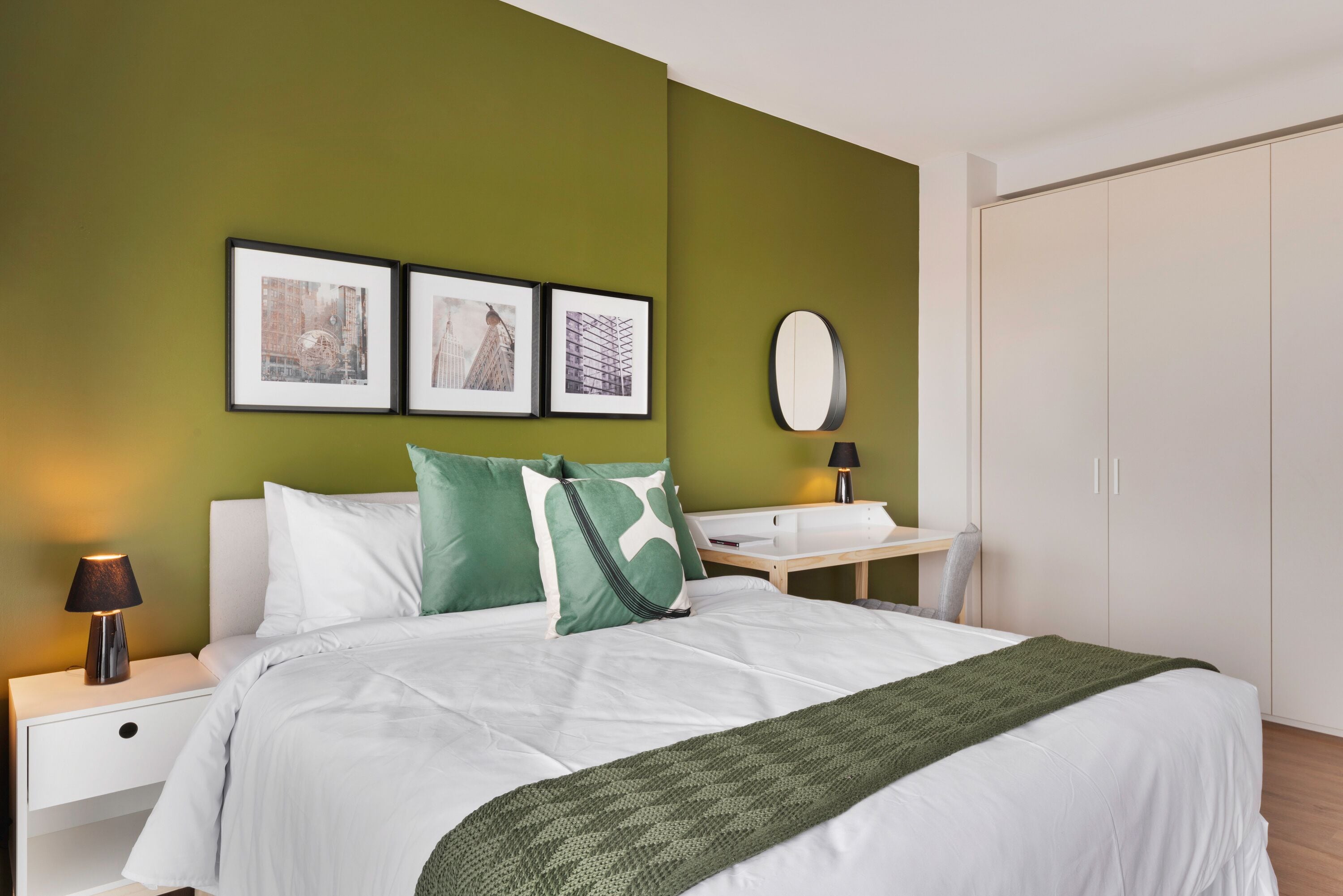 A hotel room with a bed, green walls, framed pictures, a bedside table with a lamp, and a wardrobe.