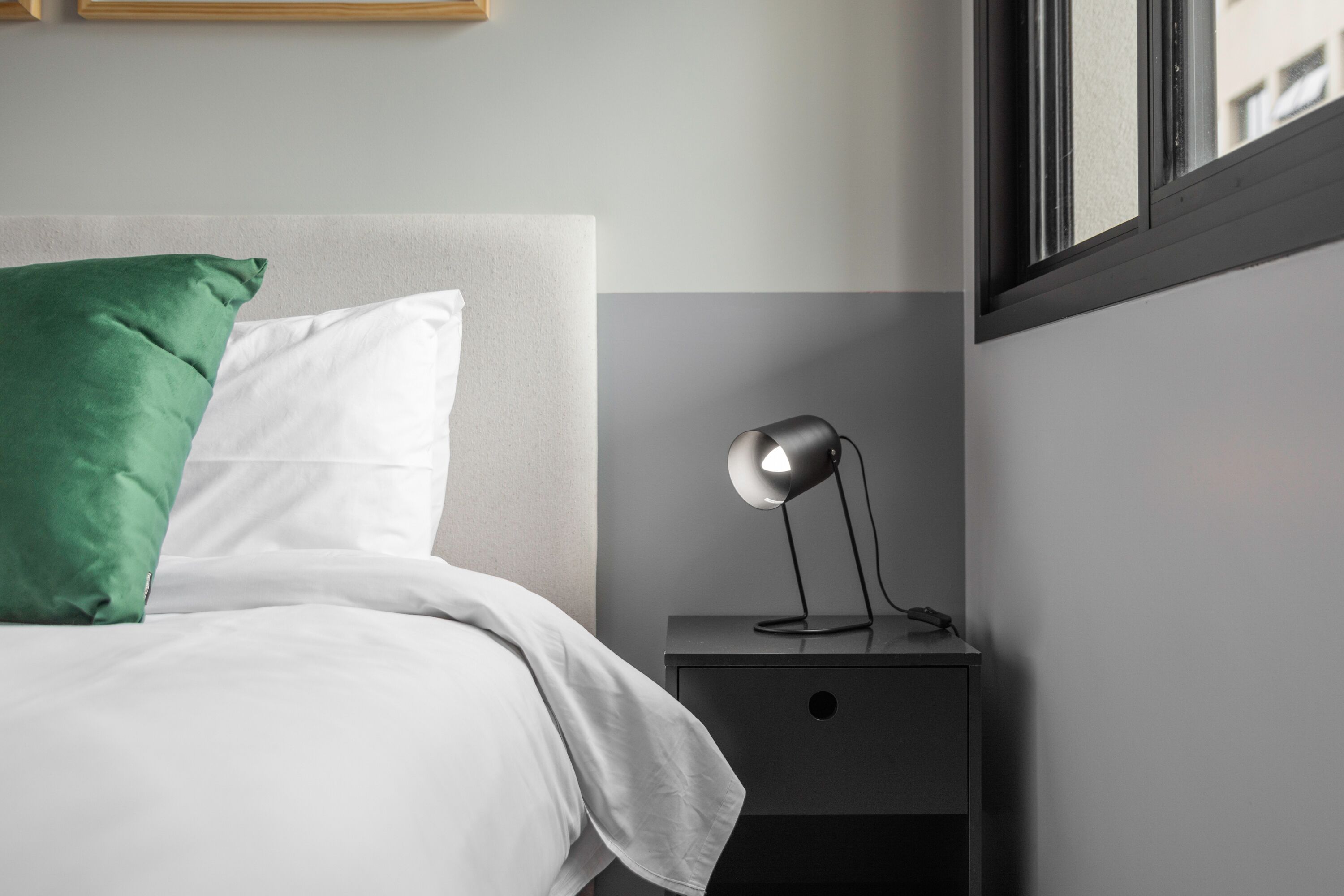 A neatly made bed with white and green pillows, a bedside table with a lamp, and a window with a view of buildings.