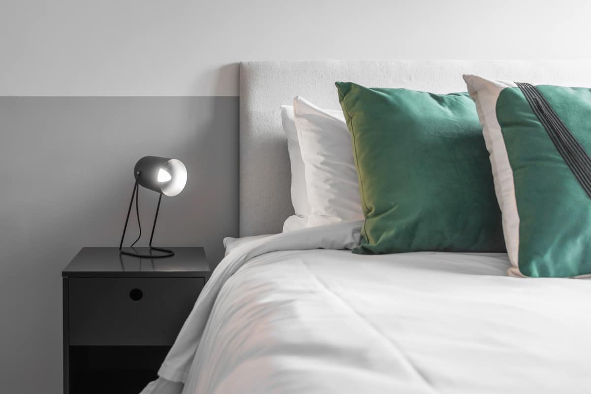 A neatly made bed with white bedding and green pillows, a bedside lamp on a black nightstand, and a grey headboard.