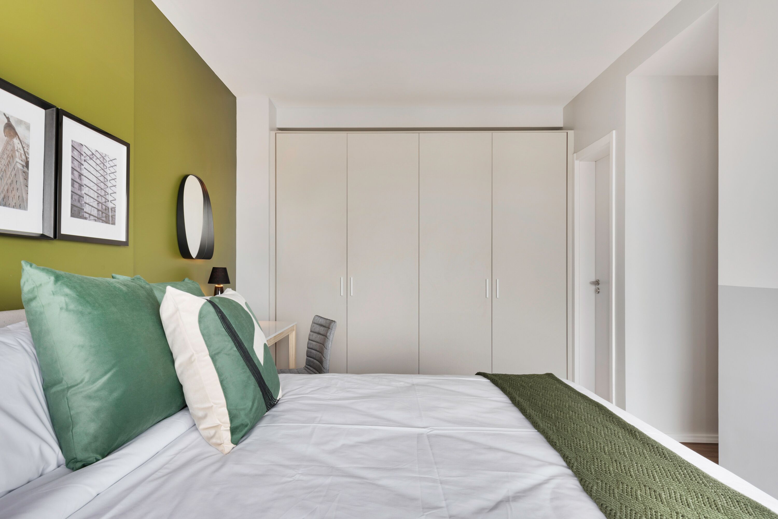 Ein Schlafzimmer mit einem Bett, grün-weißen Kissen, einem Nachttisch mit Lampe, einem Spiegel und einem Kleiderschrank.