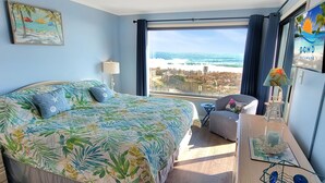 Um quarto com uma cama grande, vista para o oceano, uma mesinha com abajur e uma cadeira.