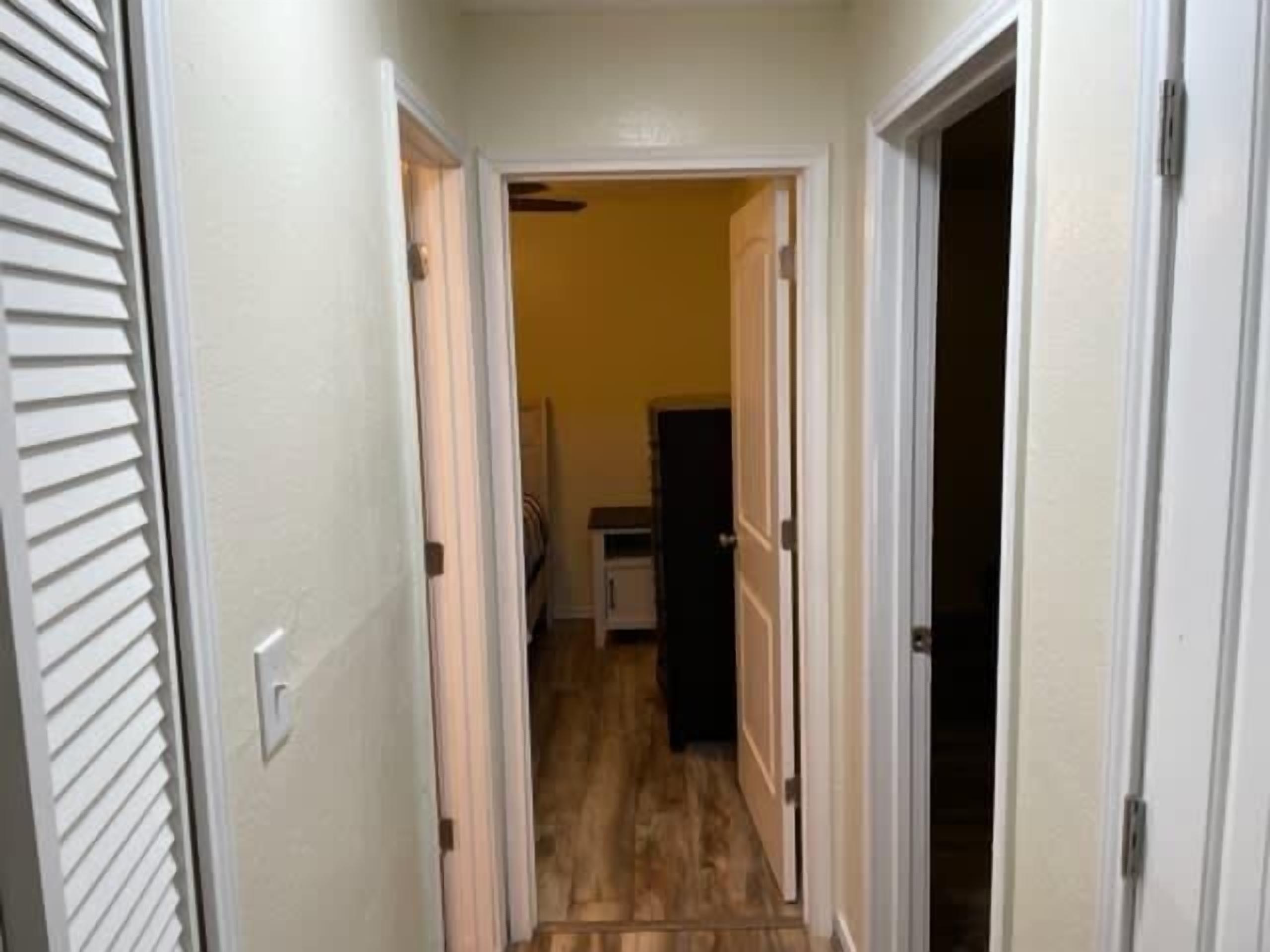 A narrow hallway with a door on the left and a door on the right, leading to a room with a visible refrigerator.