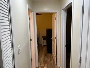 Un couloir étroit, avec une porte à gauche et une porte à droite, qui mène à une pièce où l’on peut voir un réfrigérateur.