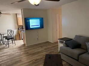 A living room with a flat-screen TV mounted on the wall, a sofa, a coffee table, and a dining area with chairs. - Comfortable downstairs 2 bed next to Fort Sill! (Lawton)