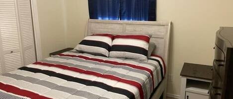 Una cama con cubrecama a rayas, una mesita de noche con lámpara y una ventana con cortinas.