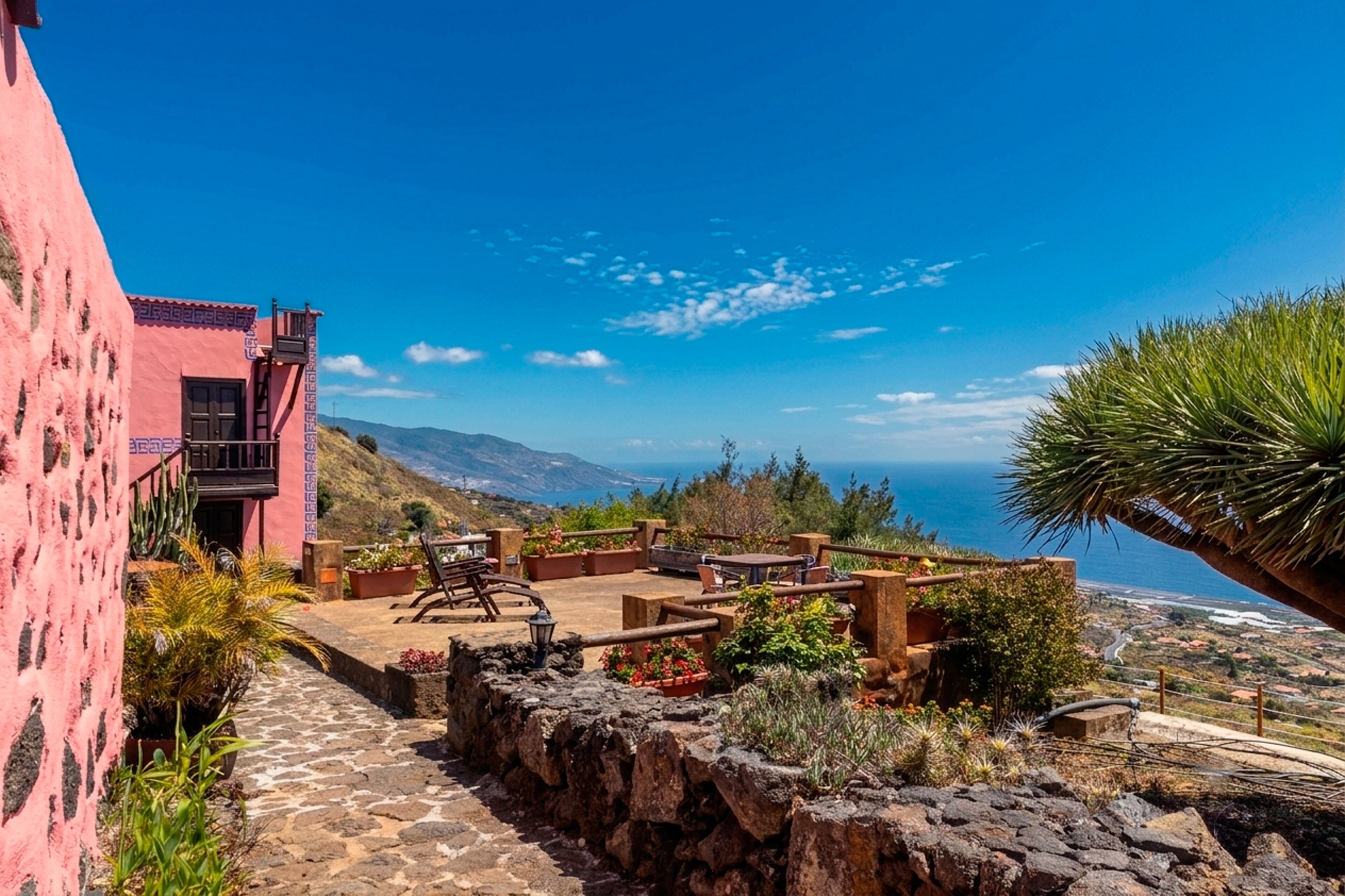 A pink building with a patio area featuring outdoor furniture and plants, overlooking a coastal landscape.