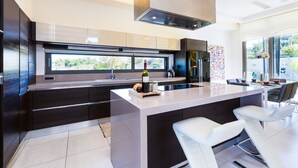A modern kitchen with dark cabinetry, a large island with bar stools, a built-in oven, and a window providing a view of the outdoors. - Lux Villa 600mt to beach★Gym & Basketball ★Heated pool★100 mt to Taverna (Platanias)