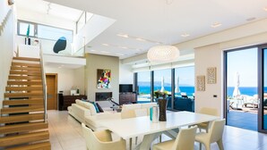 A modern living room with a dining area, a staircase, and a large window overlooking the sea. - Lux Villa 600mt to beach★Gym & Basketball ★Heated pool★100 mt to Taverna (Platanias)