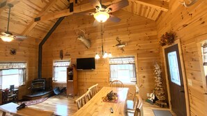 Een gezellig chaletinterieur met een houten eettafel, stoelen, een open haard en een plafondventilator.