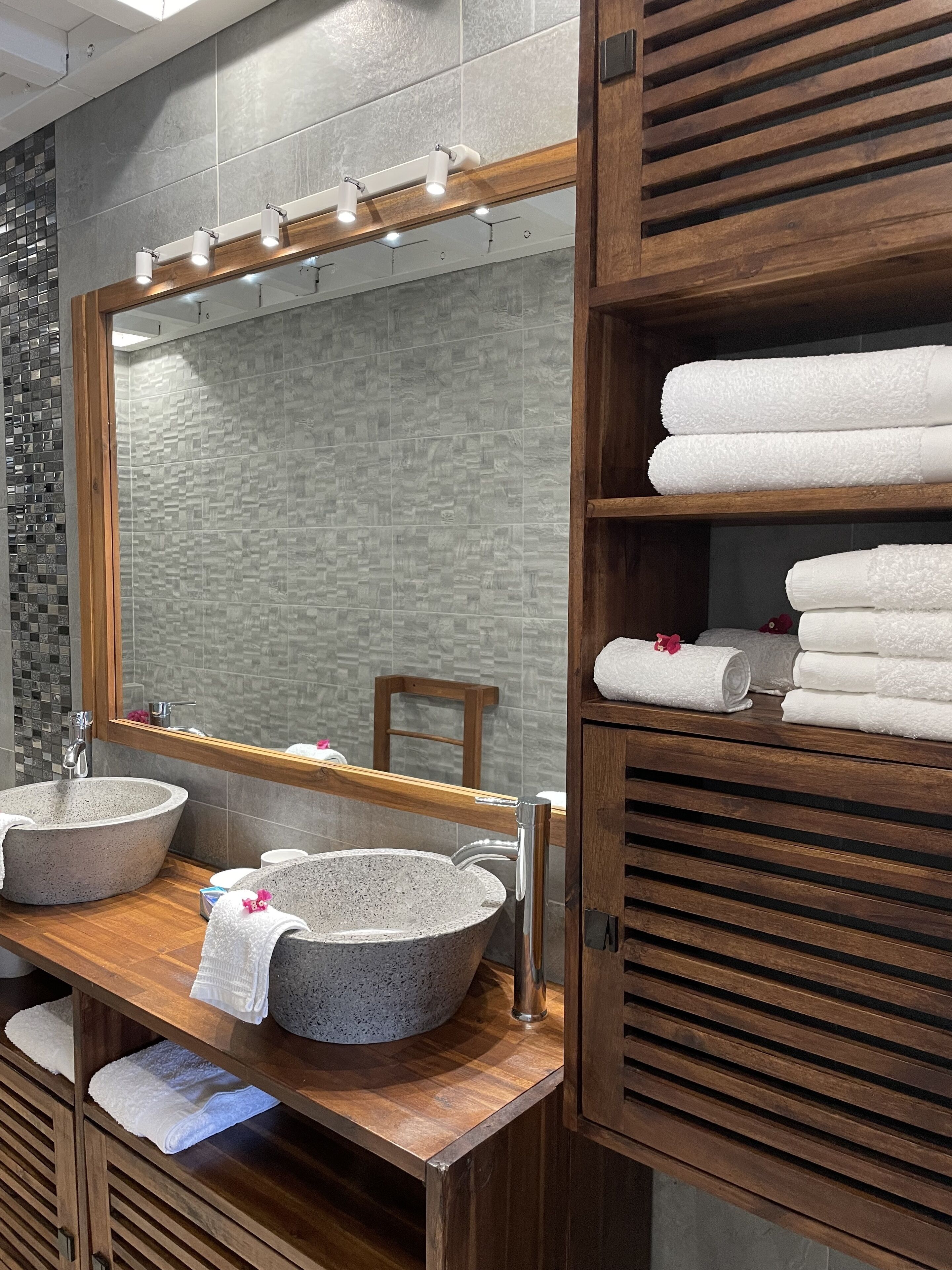 Un baño con tocador de madera, dos lavamanos de piedra, un espejo grande con iluminación y un toallero con toallas dobladas.
