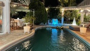 A swimming pool with lounge chairs and tables, surrounded by greenery and a covered patio area.