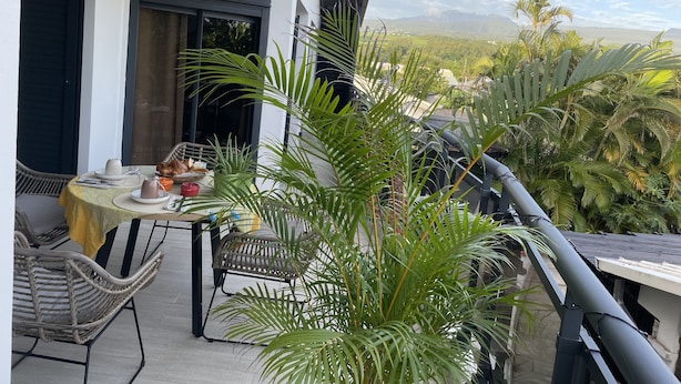A balcony with a table set for two, surrounded by wicker chairs, a potted plant, and a view of a lush landscape.