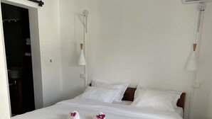 A neatly made double bed with white linens and two towels with pink flowers, a bedside table with a lamp, and an air conditioning unit on the wall.