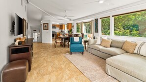 A spacious living area with a large sectional sofa, a blue armchair, and a brown ottoman. A flat-screen TV is mounted on the wall, and there is a ceiling fan.