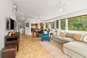 A spacious living area with a large sectional sofa, a blue armchair, and a brown ottoman. A flat-screen TV is mounted on the wall, and there is a ceiling fan.