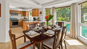 A dining area with a wooden table set for four, a kitchen in the background, and a view of a swimming pool through large windows.