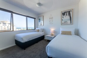 Une chambre avec deux lits, une grande fenêtre, une lampe fixée au mur et un tableau représentant une scène de plage.