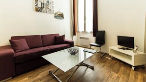 Un salon avec un canapé, une table basse en verre, un téléviseur posé sur un meuble et une fenêtre avec des rideaux.