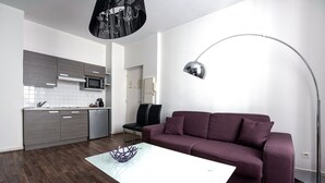 Un salon moderne avec un canapé violet, une table basse blanche et un coin cuisine doté de meubles gris et d’un four à micro-ondes.
